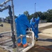 Two workers in blue hazmat/chemical protective suits and respirator masks are working on an industrial pipe system, with one leaning over the pipes and another standing nearby with one arm raised. Water or liquid is visibly spraying from the upper pipe connections, suggesting an active leak or pressurized system. The setting appears to be an outdoor industrial or municipal utility facility.