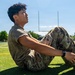 A young person wearing military camouflage uniform and tan combat boots is seated on a green artificial turf athletic field, looking upward. The setting appears to be a football field, identifiable by the yellow goal posts visible in the background. The image is taken on a clear, sunny day with trees and stadium lights visible in the distance.