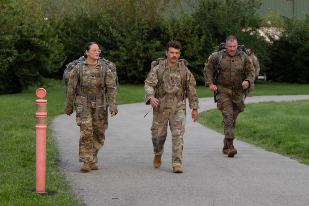 Three military personnel in camouflage uniforms (OCP/MultiCam pattern) are walking along a paved path while carrying large rucksacks on their backs. They appear to be conducting a ruck march or similar physical training exercise. Additional personnel are visible in the background on the same path.