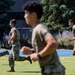 A group of military personnel in OCP (Operational Camouflage Pattern) uniforms and olive drab t-shirts are running on an athletic field with a blue track visible in the background. The setting appears to be a military installation or academy with well-maintained grounds, trees, and flowering shrubs. The image captures a physical training (PT) session in progress during daylight hours.