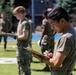 Military personnel in olive/camouflage uniforms are conducting what appears to be a physical fitness test or training evaluation on a sports field. At least two individuals in the foreground are holding clipboards and writing, suggesting they are scorers or evaluators. Additional personnel are visible in the background, including someone on the ground who may be performing an exercise.