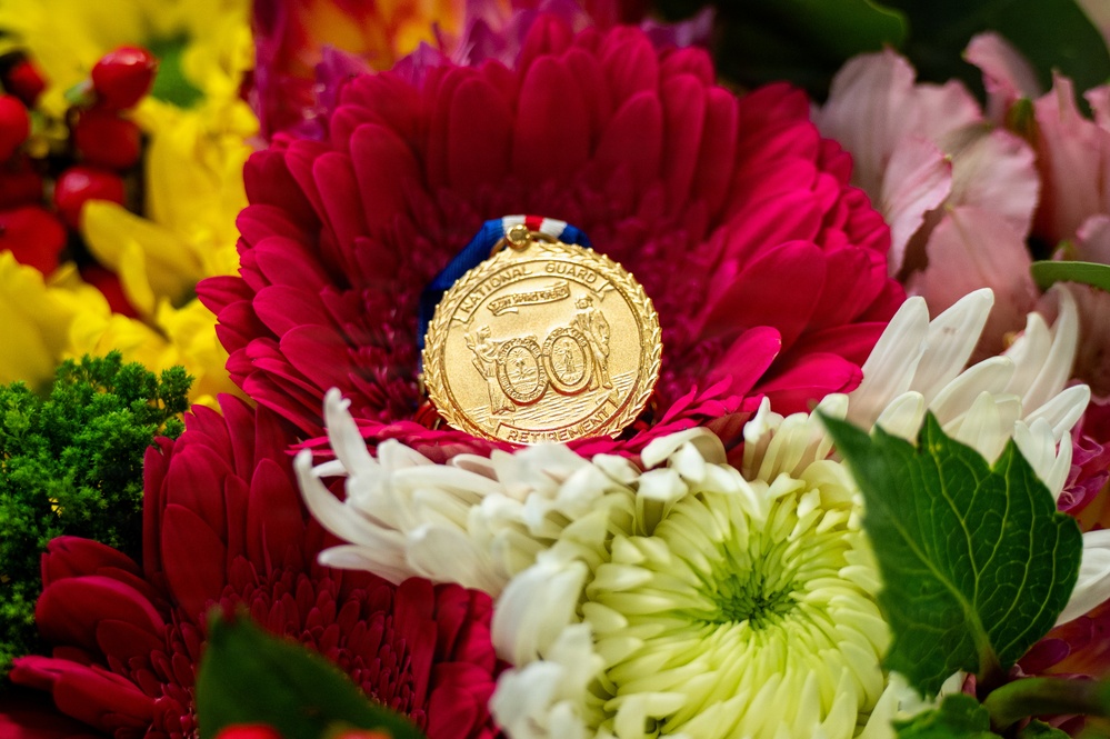 A gold National Guard Retirement medal rests on a vibrant red gerbera daisy surrounded by a colorful floral arrangement. The medal features engraved text reading 'NATIONAL GUARD' and 'RETIREMENT' with decorative imagery, suspended from a red, white, and blue ribbon. The composition suggests a retirement ceremony or commemorative occasion honoring military service.