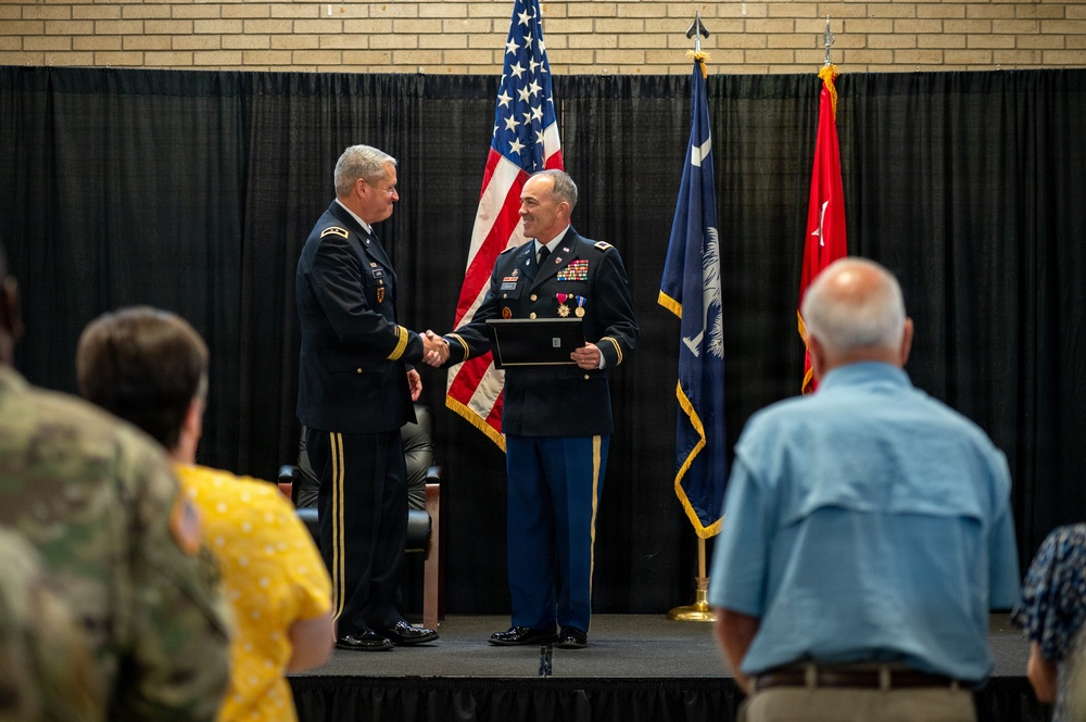 A military ceremony is taking place on a stage where two Army officers in dress uniforms are shaking hands, with one presenting a framed award or commendation to the other. The event appears to be a formal recognition or retirement ceremony, with an audience of military and civilian attendees visible in the foreground. Three flags are displayed in the background, including the U.S. flag and what appears to be a state flag.