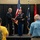 A military ceremony is taking place on a stage where two Army officers in dress uniforms are shaking hands, with one presenting a framed award or commendation to the other. The event appears to be a formal recognition or retirement ceremony, with an audience of military and civilian attendees visible in the foreground. Three flags are displayed in the background, including the U.S. flag and what appears to be a state flag.