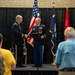 A military ceremony is taking place on a stage where two Army officers in dress uniforms are shaking hands, with one presenting a framed award or commendation to the other. The event appears to be a formal recognition or retirement ceremony, with an audience of military and civilian attendees visible in the foreground. Three flags are displayed in the background, including the U.S. flag and what appears to be a state flag.