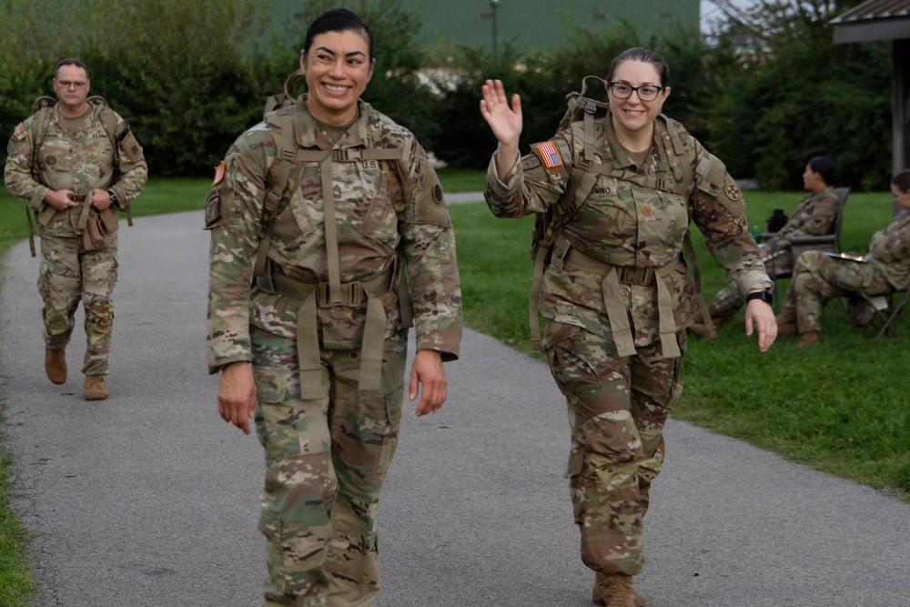 Three U.S. Army soldiers in OCP (Operational Camouflage Pattern) uniforms walk along a paved path, with two female soldiers in the foreground smiling and waving at the camera. They are wearing tactical gear including rucksacks and appear to be completing or participating in a ruck march or physical training event. Additional soldiers can be seen resting on chairs in the background.
