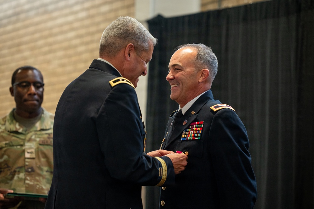 A formal U.S. Army military ceremony in which a senior officer is pinning a medal or decoration onto another officer's dress uniform. The recipient is smiling broadly, suggesting this is a positive recognition event such as a promotion or award ceremony. A third soldier in combat uniform stands in the background, appearing to hold a document or folder.