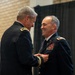 A formal U.S. Army military ceremony in which a senior officer is pinning a medal or decoration onto another officer's dress uniform. The recipient is smiling broadly, suggesting this is a positive recognition event such as a promotion or award ceremony. A third soldier in combat uniform stands in the background, appearing to hold a document or folder.