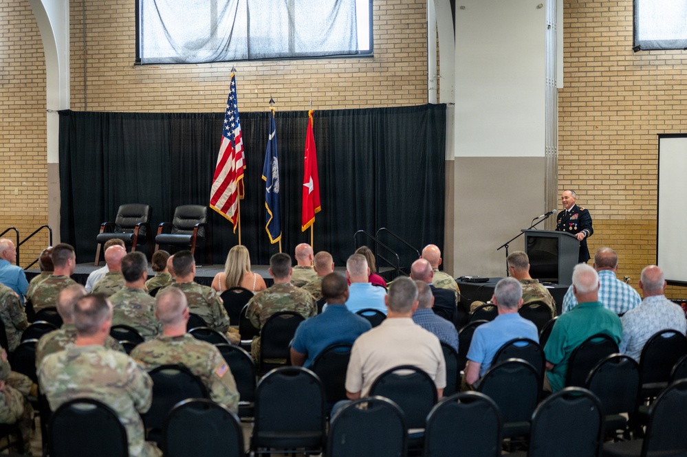 A military ceremony is taking place in a brick-walled hall where a uniformed officer is delivering a speech at a podium. The audience consists of a mix of military personnel in camouflage uniforms and civilians seated in rows of black chairs. Three flags are displayed on the stage, including the U.S. flag, a state flag, and a red general officer flag with a single star.