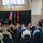 A military ceremony is taking place in a brick-walled hall where a uniformed officer is delivering a speech at a podium. The audience consists of a mix of military personnel in camouflage uniforms and civilians seated in rows of black chairs. Three flags are displayed on the stage, including the U.S. flag, a state flag, and a red general officer flag with a single star.
