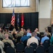 A military ceremony is taking place in a brick-walled hall where a uniformed officer is delivering a speech at a podium. The audience consists of a mix of military personnel in camouflage uniforms and civilians seated in rows of black chairs. Three flags are displayed on the stage, including the U.S. flag, a state flag, and a red general officer flag with a single star.