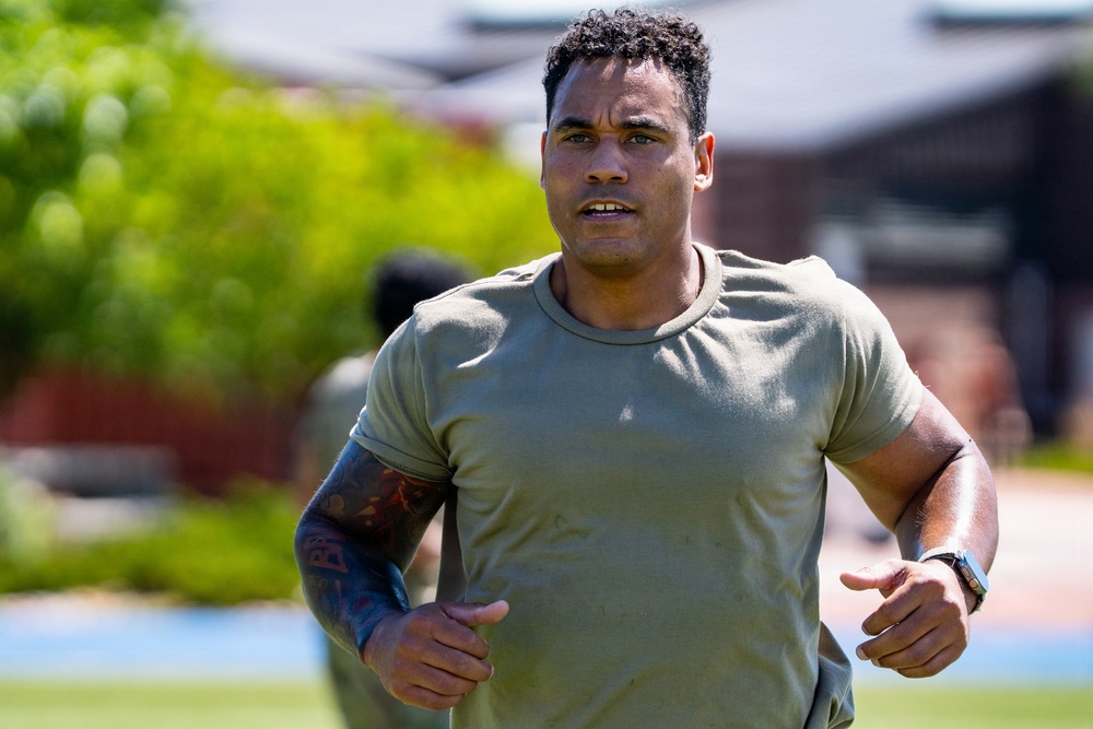 A muscular man wearing an olive/sage green military-style t-shirt is running outdoors in what appears to be a physical training exercise. He has short curly hair, a sleeve tattoo on his left arm, and is wearing a watch on his right wrist. The background shows blurred green trees and brick buildings, suggesting a military base or institutional campus setting.