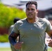 A muscular man wearing an olive/sage green military-style t-shirt is running outdoors in what appears to be a physical training exercise. He has short curly hair, a sleeve tattoo on his left arm, and is wearing a watch on his right wrist. The background shows blurred green trees and brick buildings, suggesting a military base or institutional campus setting.