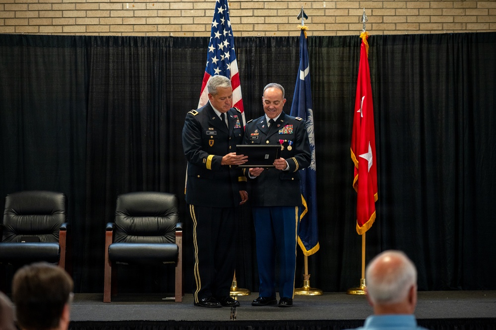 Two U.S. Army officers in dress uniforms are on a stage during a formal military ceremony, jointly holding a framed award or certificate. Behind them stand multiple flags including the U.S. flag, what appears to be a state flag, and a general officer's flag with a single star. An audience is partially visible in the foreground.