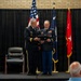 Two U.S. Army officers in dress uniforms are on a stage during a formal military ceremony, jointly holding a framed award or certificate. Behind them stand multiple flags including the U.S. flag, what appears to be a state flag, and a general officer's flag with a single star. An audience is partially visible in the foreground.