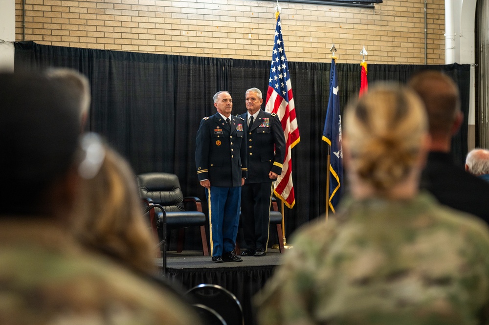 A formal U.S. Army military ceremony taking place indoors, likely a change of command or retirement ceremony. Two senior Army officers in dress blue uniforms stand at attention on a raised platform stage, flanked by the American flag and what appears to be a unit or state flag. An audience of military personnel in Army Combat Uniforms (ACU) and civilians observe from the foreground.