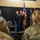 A formal U.S. Army military ceremony taking place indoors, likely a change of command or retirement ceremony. Two senior Army officers in dress blue uniforms stand at attention on a raised platform stage, flanked by the American flag and what appears to be a unit or state flag. An audience of military personnel in Army Combat Uniforms (ACU) and civilians observe from the foreground.