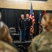 A formal U.S. Army military ceremony taking place indoors, likely a change of command or retirement ceremony. Two senior Army officers in dress blue uniforms stand at attention on a raised platform stage, flanked by the American flag and what appears to be a unit or state flag. An audience of military personnel in Army Combat Uniforms (ACU) and civilians observe from the foreground.