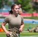 A young woman in military-style physical training attire is running or jogging on a grass field. She is wearing an olive drab t-shirt, camouflage pants, and a tan tactical belt, suggesting a military training or ROTC exercise. The background shows a sports field with other personnel and equipment visible in soft focus.