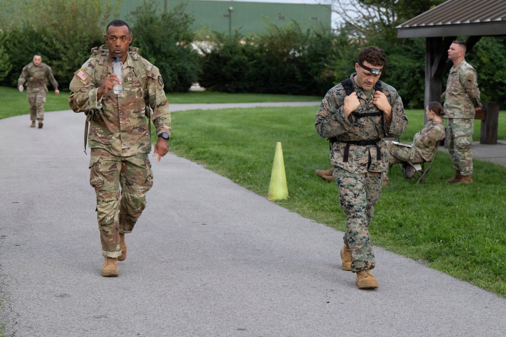 The image shows military personnel in a outdoor setting, appearing to participate in a physical training or ruck march event on a paved path. Two service members are prominently in the foreground — one wearing U.S. Army OCP (Operational Camouflage Pattern) uniform and one wearing U.S. Marine Corps MARPAT uniform, both carrying backpacks. Additional soldiers are visible in the background, including observers near a covered pavilion.