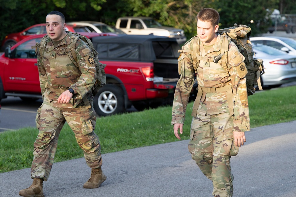 Two U.S. Army soldiers in Operational Camouflage Pattern (OCP) uniforms are walking along a paved path while carrying rucksacks on their backs. Both soldiers appear to be conducting a ruck march or movement exercise in what appears to be a military installation or base area. A parking lot with civilian vehicles is visible in the background.