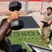 A United States Air Force physical fitness assessment is taking place on an outdoor turf area marked with the Air Force logo and text. One service member in a t-shirt is performing a dumbbell exercise while another uniformed service member holds a clipboard, apparently recording scores or repetitions. A third uniformed individual is partially visible on the right edge of the frame.