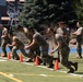 A group of military personnel in camouflage uniforms are performing an agility drill on an artificial turf athletic field, navigating through a line of orange traffic cones. Several participants are in a low crouching stance moving laterally along the cone line, while two individuals in the background appear to be observing and recording results. The setting appears to be a military physical fitness assessment or training exercise conducted outdoors during daytime.