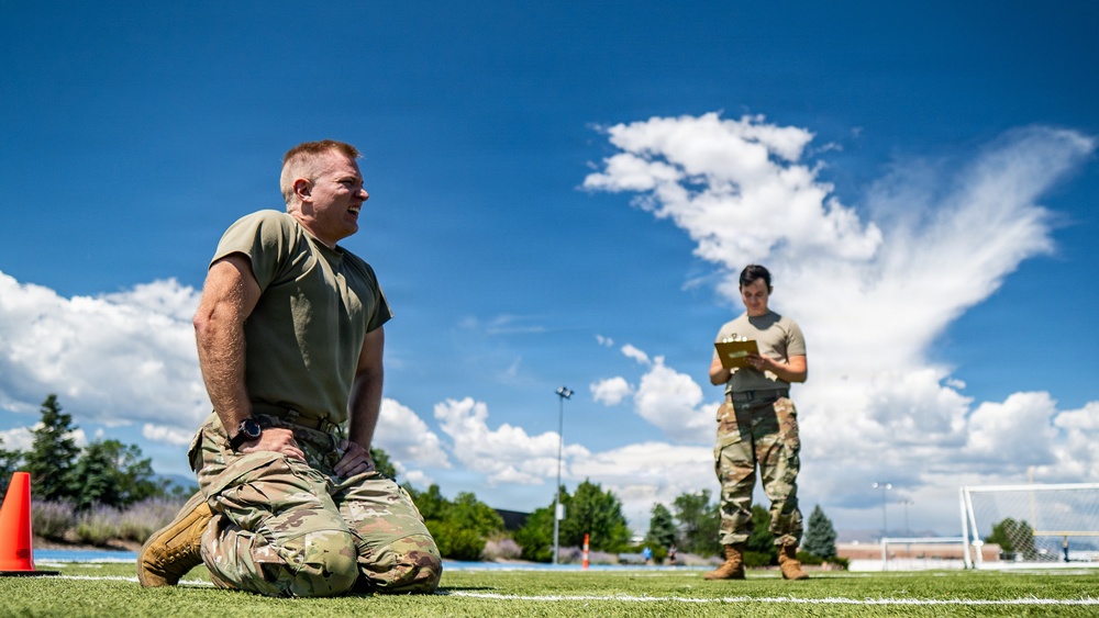 Two military personnel are on an outdoor athletic field, likely conducting a physical fitness test or assessment. The foreground soldier is kneeling on the turf in a resting position after exertion, while a second soldier in the background records data on a clipboard. The setting appears to be a military installation sports field under bright, partly cloudy skies.
