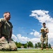 Two military personnel are on an outdoor athletic field, likely conducting a physical fitness test or assessment. The foreground soldier is kneeling on the turf in a resting position after exertion, while a second soldier in the background records data on a clipboard. The setting appears to be a military installation sports field under bright, partly cloudy skies.