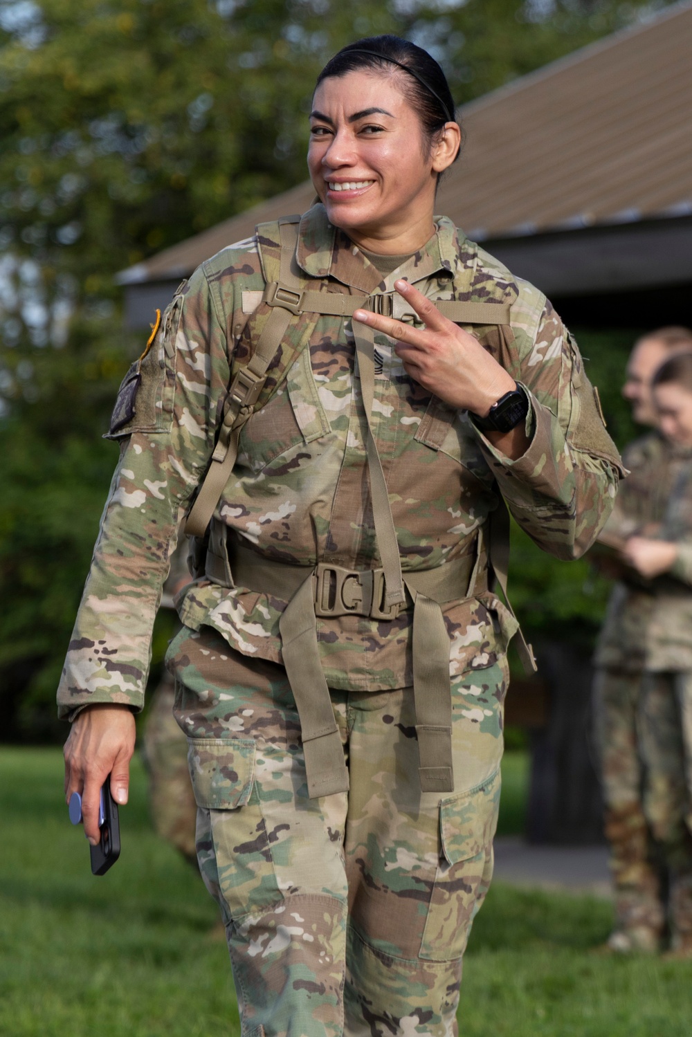 A female soldier in US Army OCP (Operational Camouflage Pattern) uniform is smiling and making a peace/victory sign with her right hand. She is wearing a tactical harness/ruck sack straps and a black wristwatch, holding what appears to be a small device in her left hand. The setting appears to be an outdoor military training or garrison environment.