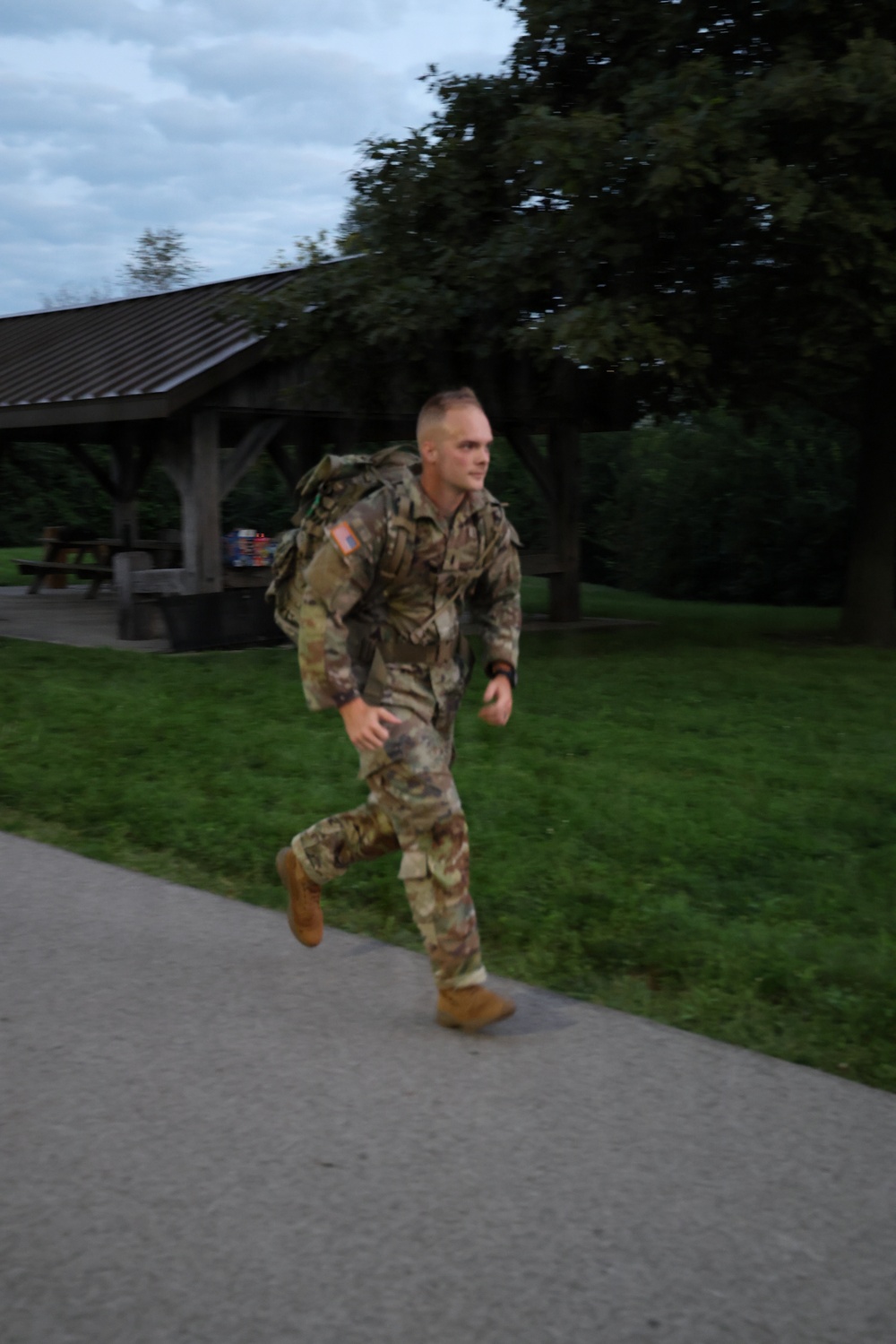 A male soldier in US Army OCP (Operational Camouflage Pattern) uniform is running along a paved path while carrying a military rucksack. He appears to be conducting a ruck march or physical training exercise in what looks like a park or recreational area. The image was taken in low-light conditions, likely early morning or evening.
