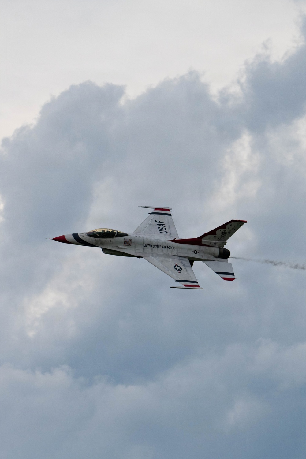 A United States Air Force Thunderbirds F-16 Fighting Falcon is captured in flight against a partly cloudy sky. The aircraft displays the distinctive Thunderbirds livery of white with red, white, and blue markings. A faint smoke trail is visible behind the aircraft, suggesting a demonstration maneuver.