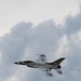 A United States Air Force Thunderbirds F-16 Fighting Falcon is captured in flight against a partly cloudy sky. The aircraft displays the distinctive Thunderbirds livery of white with red, white, and blue markings. A faint smoke trail is visible behind the aircraft, suggesting a demonstration maneuver.