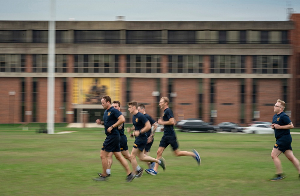 A group of individuals in matching dark navy blue athletic uniforms with gold lettering are running across a grass field. They appear to be military or naval personnel conducting a physical training exercise in front of a large multi-story brick building. The image uses motion blur to convey speed and movement.