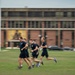 A group of individuals in matching dark navy blue athletic uniforms with gold lettering are running across a grass field. They appear to be military or naval personnel conducting a physical training exercise in front of a large multi-story brick building. The image uses motion blur to convey speed and movement.