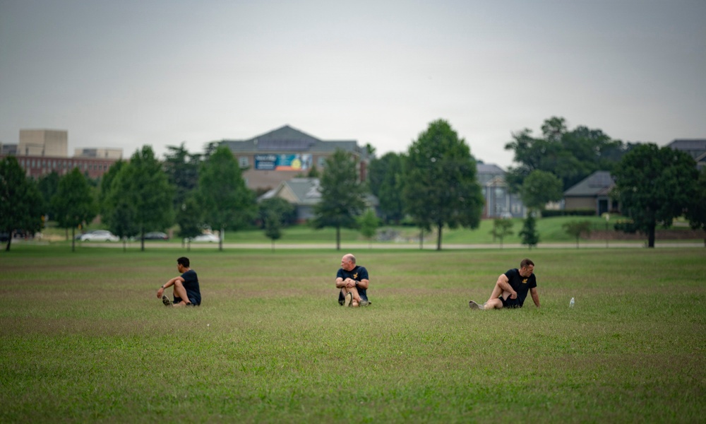 Three individuals are seated on a large open grass field, appearing to stretch or rest after physical activity. They are spaced apart from one another and are wearing matching dark athletic clothing. A backdrop of trees and institutional buildings is visible in the soft-focus background.