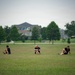 Three individuals are seated on a large open grass field, appearing to stretch or rest after physical activity. They are spaced apart from one another and are wearing matching dark athletic clothing. A backdrop of trees and institutional buildings is visible in the soft-focus background.