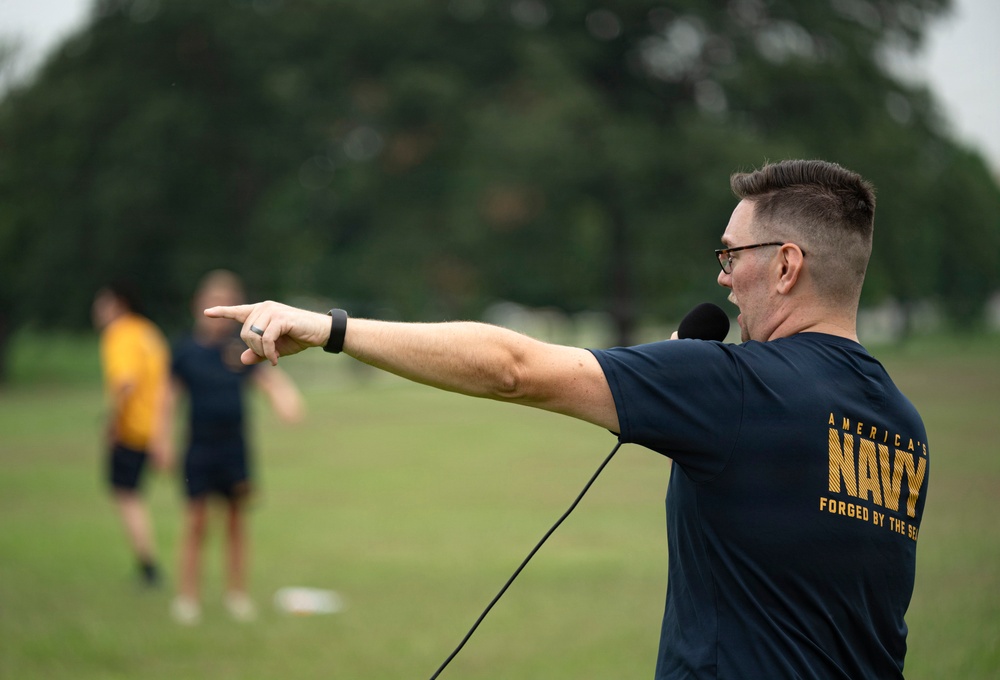 A man wearing an 'America's Navy - Forged by the Sea' t-shirt is pointing directions on an outdoor field while holding a microphone or communication device. He appears to be in an instructional or leadership role, directing personnel visible in the blurred background. The setting appears to be a physical training or outdoor exercise event on a grassy field.