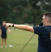 A man wearing an 'America's Navy - Forged by the Sea' t-shirt is pointing directions on an outdoor field while holding a microphone or communication device. He appears to be in an instructional or leadership role, directing personnel visible in the blurred background. The setting appears to be a physical training or outdoor exercise event on a grassy field.