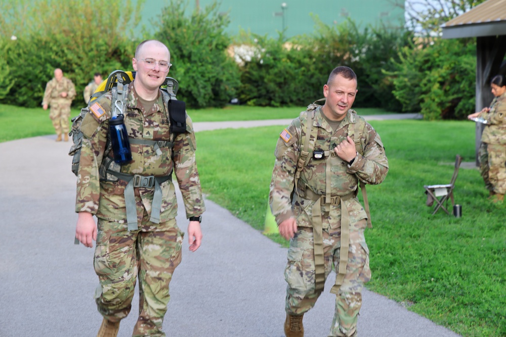 Two U.S. Army soldiers in Operational Camouflage Pattern (OCP) uniforms walk along a paved path, both carrying rucksacks and wearing load-bearing equipment. The soldier on the left is smiling and wearing glasses, with a blue water bottle attached to his gear. Additional soldiers are visible in the background near a grassy area, suggesting a unit physical training or ruck march event.