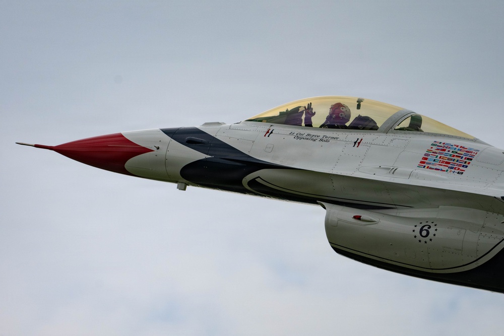 A close-up photograph of a U.S. Air Force Thunderbirds F-16 Fighting Falcon, numbered '6', in flight against an overcast sky. The aircraft displays the distinctive Thunderbirds red, white, and blue livery, and the pilot is visible in the cockpit appearing to wave. Text on the fuselage reads 'Lt. Col. Bryce Turner, Opposing Solo', identifying the assigned pilot of this aircraft.