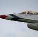 A close-up photograph of a U.S. Air Force Thunderbirds F-16 Fighting Falcon, numbered '6', in flight against an overcast sky. The aircraft displays the distinctive Thunderbirds red, white, and blue livery, and the pilot is visible in the cockpit appearing to wave. Text on the fuselage reads 'Lt. Col. Bryce Turner, Opposing Solo', identifying the assigned pilot of this aircraft.
