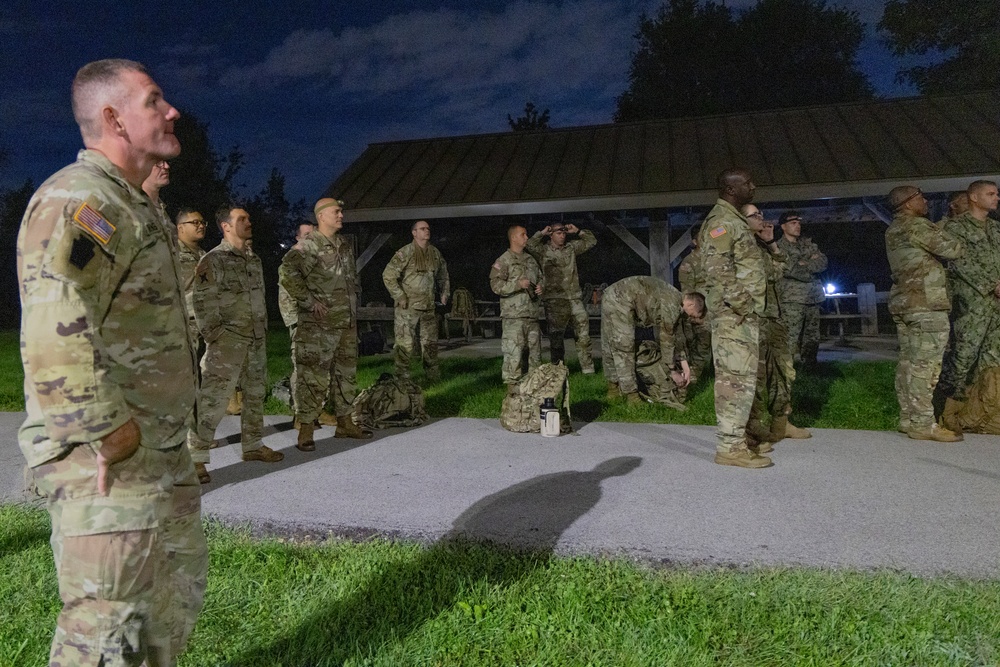 A group of U.S. Army soldiers in OCP (Operational Camouflage Pattern) uniforms are assembled outdoors at night near a covered pavilion structure. The soldiers appear to be gathered for a briefing, formation, or pre-mission assembly, with some standing at ease and others adjusting gear. The setting appears to be a military installation or training area.