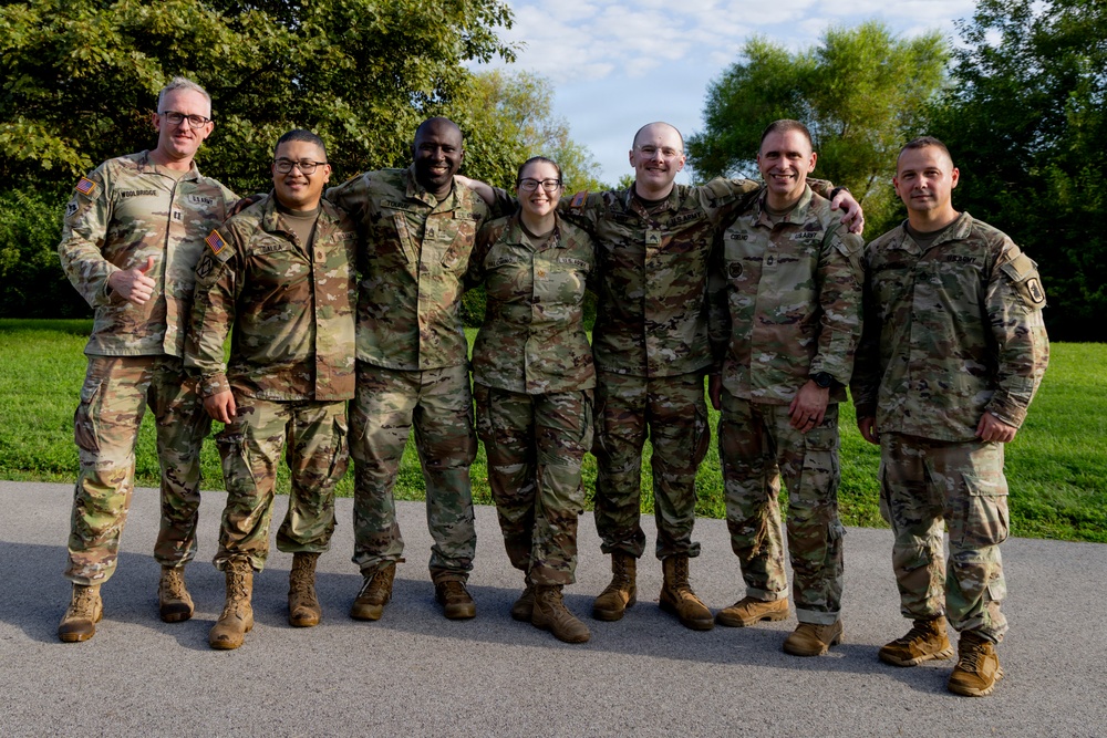 Seven U.S. Army soldiers in OCP (Operational Camouflage Pattern) uniforms pose together for a group photo outdoors. They are standing on a paved surface with a green lawn and trees visible in the background. The group includes both male and female soldiers, and several are smiling with arms around each other in a relaxed, celebratory manner.