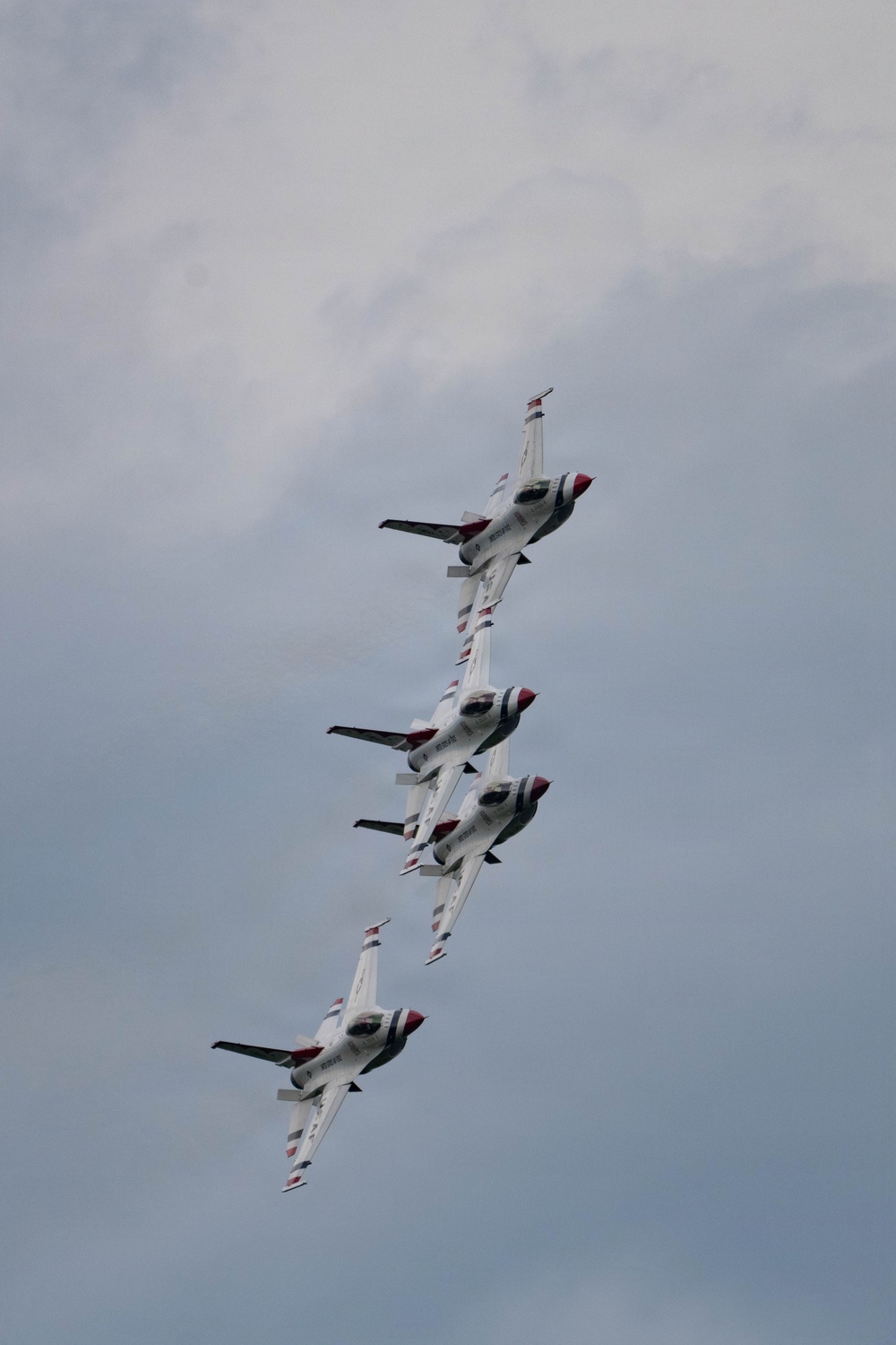 Four military jet aircraft are flying in a tight diagonal formation against an overcast sky. The aircraft display distinctive white, red, and blue livery consistent with the U.S. Air Force Thunderbirds aerial demonstration team. The jets appear to be performing a precision formation maneuver, stacked closely in a line.