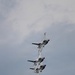 Four military jet aircraft are flying in a tight diagonal formation against an overcast sky. The aircraft display distinctive white, red, and blue livery consistent with the U.S. Air Force Thunderbirds aerial demonstration team. The jets appear to be performing a precision formation maneuver, stacked closely in a line.