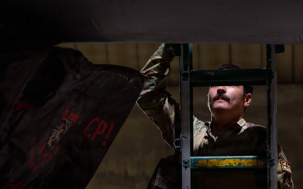 A military personnel member in camouflage uniform is standing on a green metal ladder, lifting or removing a dark cover or tarp from what appears to be military equipment or an aircraft. The image is dramatically lit with strong contrast between light and shadow, giving it a cinematic quality. Text markings including 'CP!' in red are visible on the dark cover being handled.
