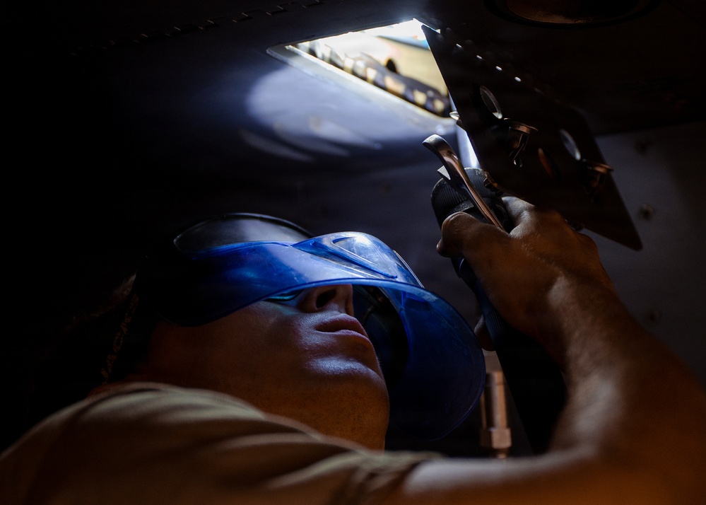 A technician or mechanic is working underneath a large vehicle or aircraft, looking upward while using a power tool or hand tool on a mechanical component. The worker is wearing blue safety goggles/glasses for eye protection. The image is taken from a low angle, creating a dramatic perspective that emphasizes the confined working space.