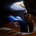 A technician or mechanic is working underneath a large vehicle or aircraft, looking upward while using a power tool or hand tool on a mechanical component. The worker is wearing blue safety goggles/glasses for eye protection. The image is taken from a low angle, creating a dramatic perspective that emphasizes the confined working space.