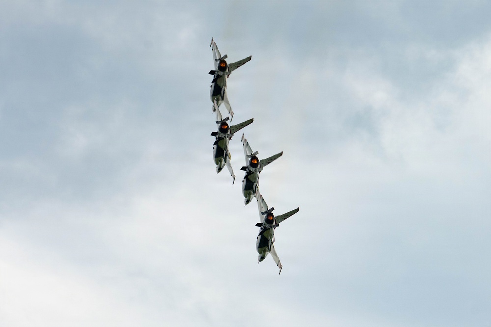 Four military jet aircraft are flying in a tight diagonal formation against an overcast sky. The jets appear to be performing an aerobatic display, flying in a stacked line-astern formation at a steep bank angle. Their afterburners appear to be engaged, visible as orange glows at the engine exhausts.